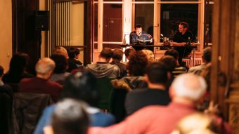 Zwei Personen auf einem Podium lesen aus einem Buch für ein Publikum vor