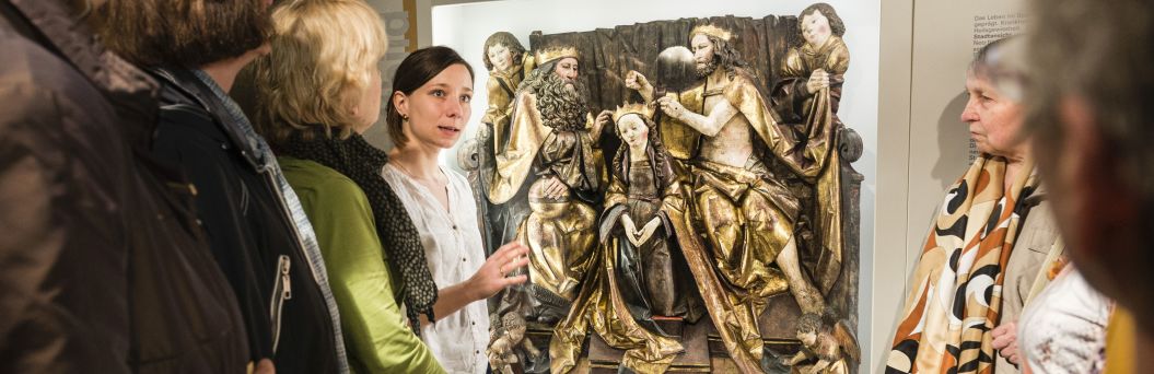 Eine Museumsführerin steht mit mehreren Menschen vor einem historischen Kunstwerk.