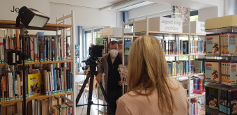 Die Fotos zeigen Impressionen vom Filmdreh in der Ernst-Abbe-Bücherei. Zu sehen ist ein Kameramann, der zwischen den Bücherregalen ein Interview führt.
